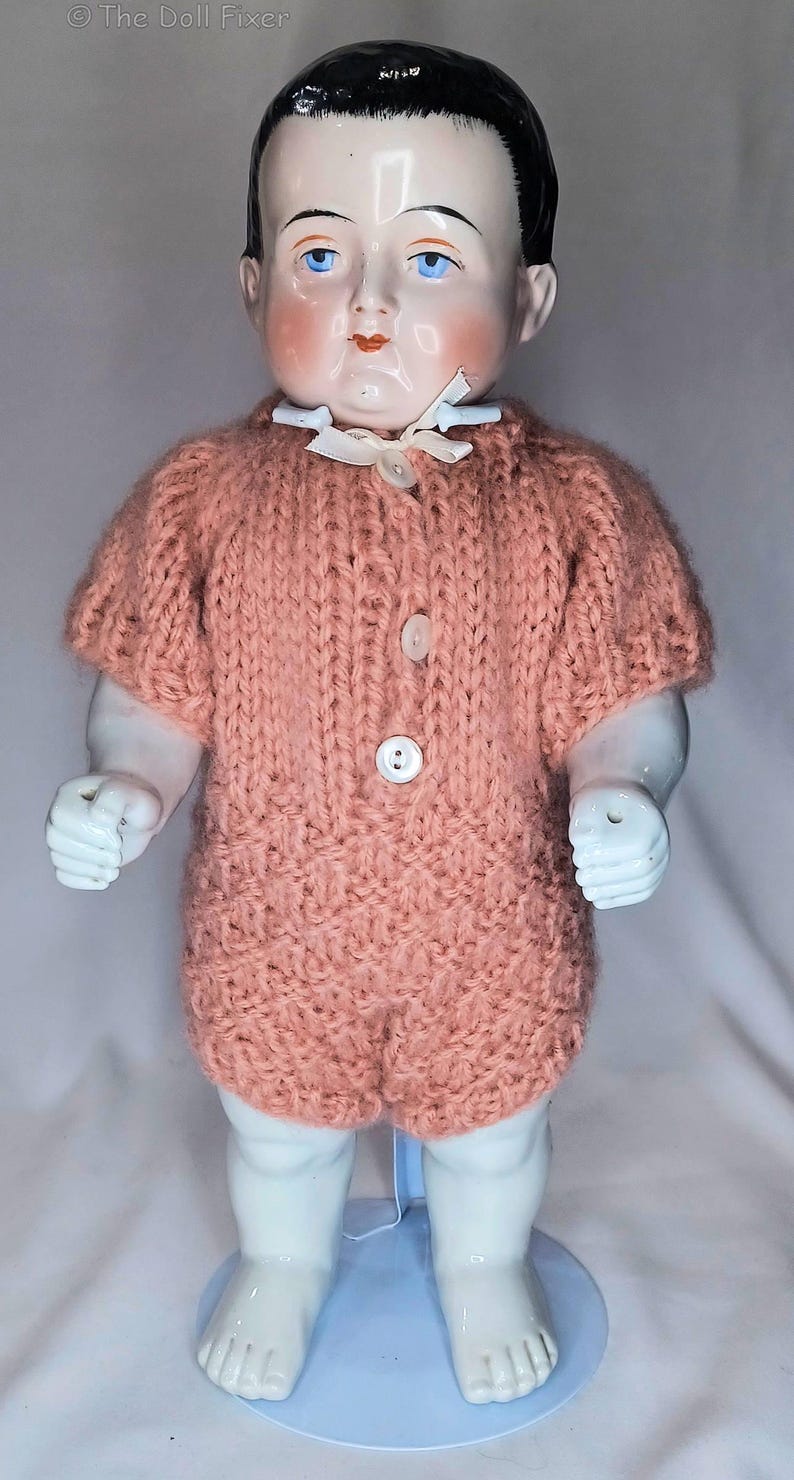 May include: A porcelain doll with painted black hair and blue eyes, wearing a short-sleeved, peach-colored knitted romper. The romper features a textured stitch pattern, a small white ribbon at the collar, and three white buttons down the front.
