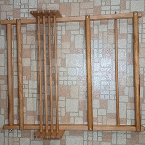 May include: A light brown wooden weaving loom frame featuring a rectangular structure with multiple vertical dowels and horizontal support bars. The frame includes adjustable metal hardware and wing nuts at the corners, with the text The Doll Fixer visible in the top left corner.