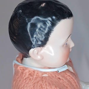 May include: A porcelain doll head with painted black hair and blue eyes, featuring a smooth, glossy finish. The doll wears a peach-colored knitted sweater with a light blue plastic collar. The text "The Doll Fixer" is visible in the top left corner.