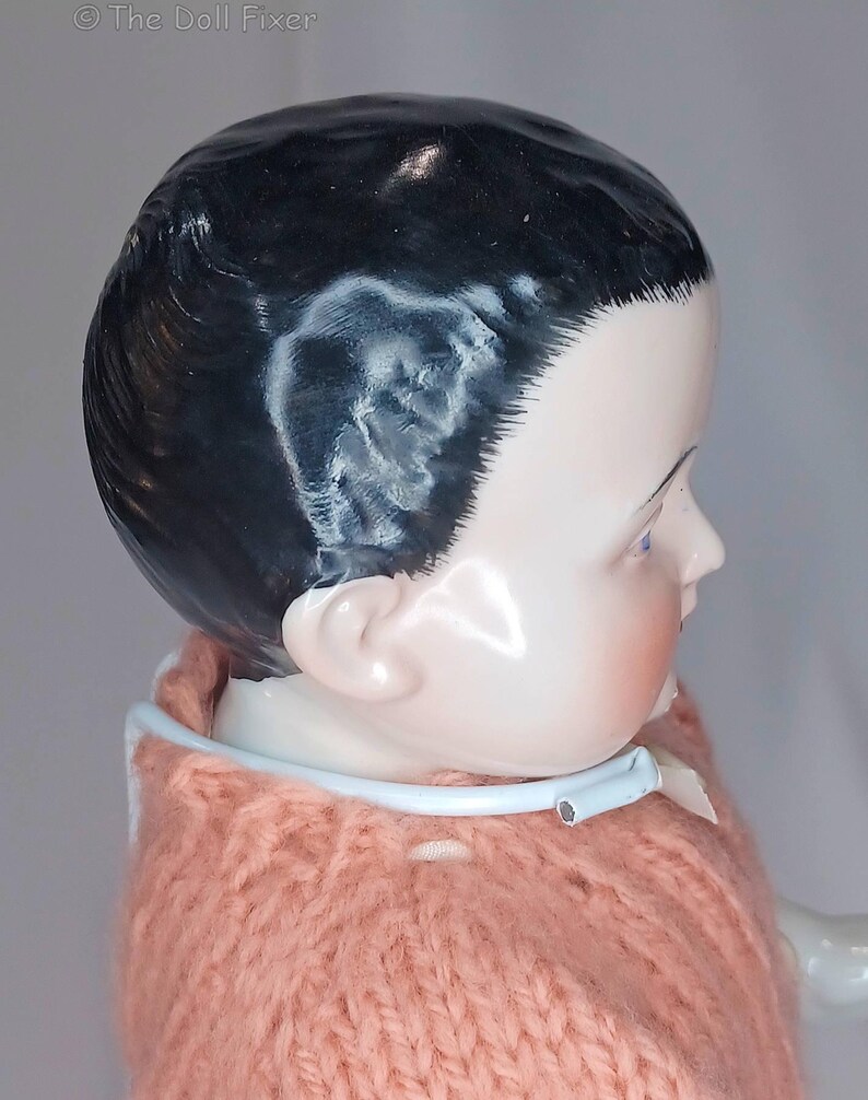 May include: A porcelain doll head with painted black hair and blue eyes, featuring a smooth, glossy finish. The doll wears a peach-colored knitted sweater with a light blue plastic collar. The text "The Doll Fixer" is visible in the top left corner.