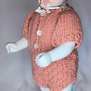 May include: A porcelain doll with black hair and blue eyes wearing a short-sleeved, peach-colored knitted romper. The romper features a round neckline with a small white ribbon bow and three white buttons down the front. The doll stands on a light blue base.