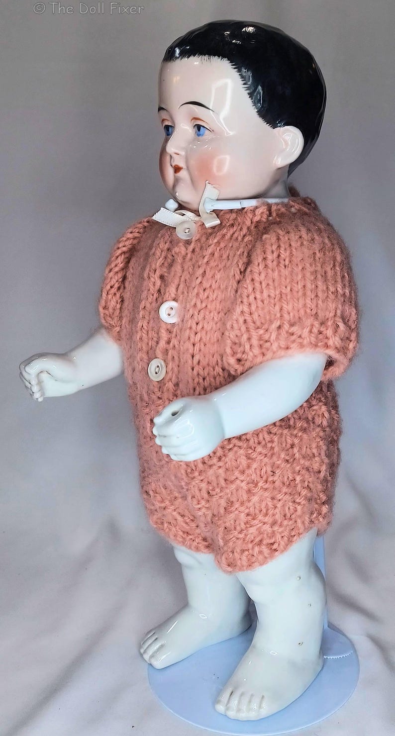 May include: A porcelain doll with black hair and blue eyes wearing a short-sleeved, peach-colored knitted romper. The romper features a round neckline with a small white ribbon bow and three white buttons down the front. The doll stands on a light blue base.