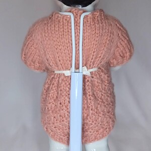 May include: A doll with black hair wearing a short-sleeved, dusty pink knitted romper. The doll is supported by a white metal stand that extends up the back, featuring a thin white ribbon tied at the waist. The text The Doll Fixer is visible.