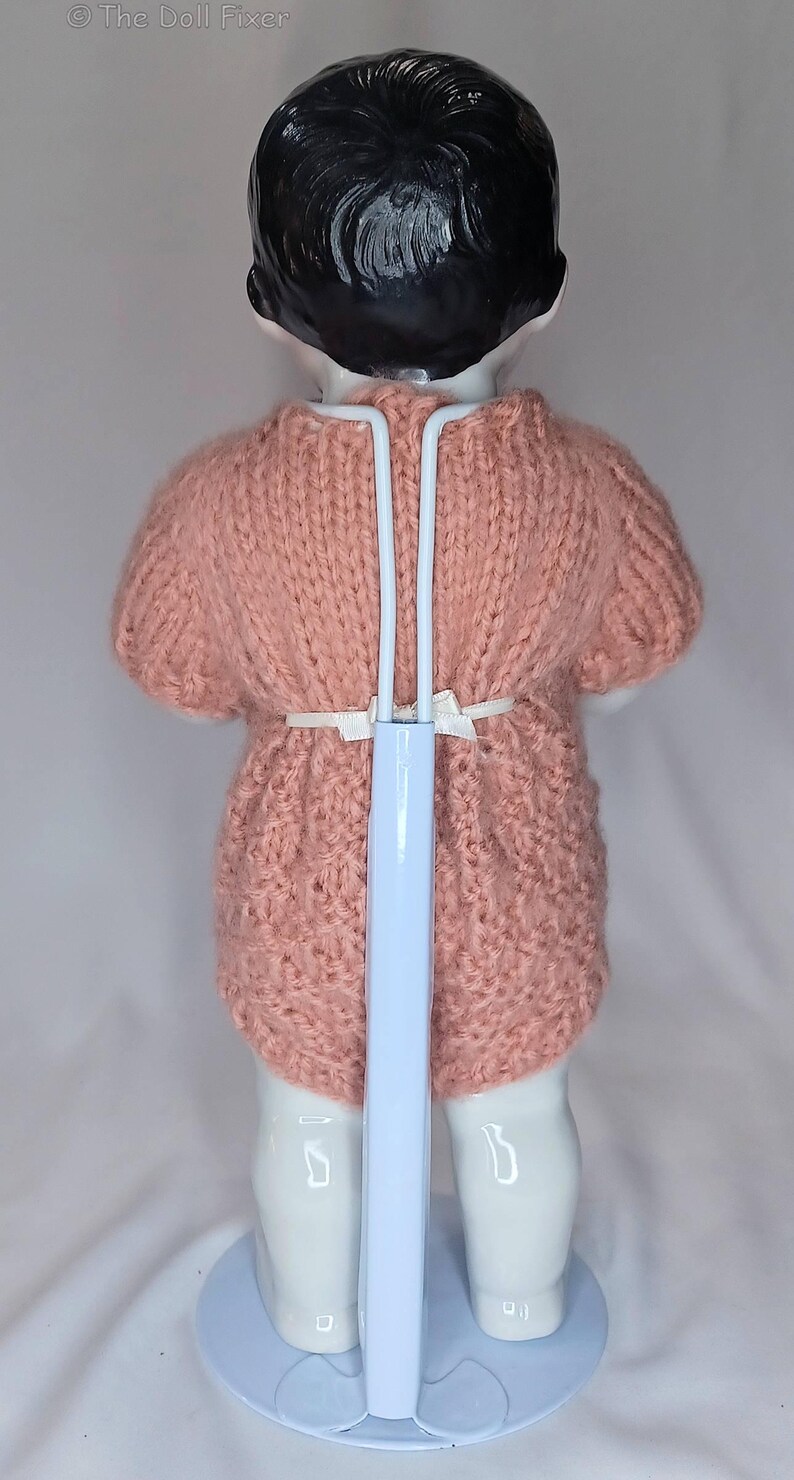 May include: A doll with black hair wearing a short-sleeved, dusty pink knitted romper. The doll is supported by a white metal stand that extends up the back, featuring a thin white ribbon tied at the waist. The text The Doll Fixer is visible.