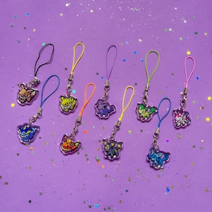 May include: A selection of pixelated character charms in various colours, including orange, yellow, blue, purple, and green. Each charm is attached to a coloured loop and silver chain, set against a purple background with glitter.