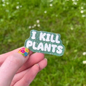 May include: A green sticker with the text "I KILL PLANTS" in white, bubble letters. The sticker has a light green border and a pattern of small white stars. The background is blurred green grass.