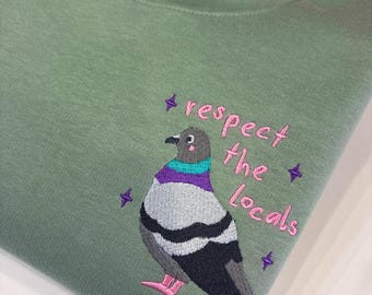 May include: A sage green crewneck sweatshirt featuring an embroidered pigeon with a purple and teal neck, grey and black wings, and pink feet. The text "respect the locals" is embroidered in pink next to the pigeon, surrounded by small purple stars.