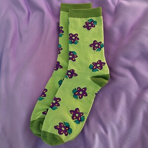 May include: A pair of green socks with a repeating pattern of purple flowers and green leaves. The socks have green cuffs, heels, and toes. The socks are displayed on a purple fabric background.
