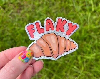 May include: A sticker with a croissant illustration and the word "FLAKY" in red bubble letters. The sticker has a silver glitter border. The background is green grass.