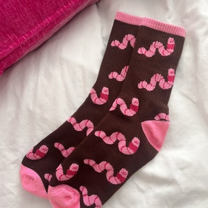 May include: A pair of brown socks featuring a repeating pattern of pink worms. The socks have pink heels and cuffs. The socks are displayed on a white surface.
