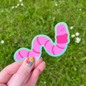 May include: A pink worm sticker with a light green border and a smiling face. The worm has a pink scarf and red stripes. The sticker is held in a hand with a floral nail design, set against a backdrop of green grass.