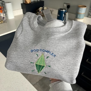 May include: A gray sweatshirt with the embroidered text "GOD COMPLEX" in blue, and a green diamond graphic. The sweatshirt is folded, with the tag visible. The garment is made of a soft material.