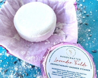 May include: A lavender-scented shower steamer in a pink and white wrapper. The steamer is round and white with the text "HANDCRAFTED Lavender Fields AROMATHERAPY SHOWER STEAMER" printed on the wrapper. The text "Net Wt. 3 oz (85g)" is also printed on the wrapper.