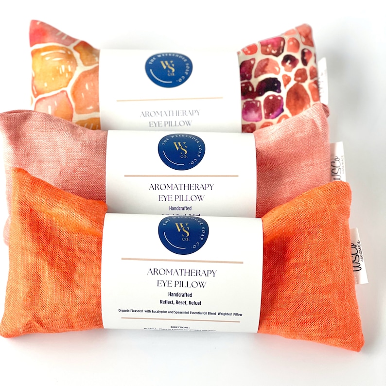 May include: Three aromatherapy eye pillows with different colored linen covers. The pillows are labeled "Aromatherapy Eye Pillow" and "Handcrafted". The top pillow has a watercolor design of orange and pink stones. The middle pillow is a solid pink color. The bottom pillow is a solid orange color.