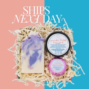 May include: A gift box filled with lavender-themed bath products. The box contains a bar of lavender soap, a jar of lavender sugar scrub, and a lavender bath bomb. The box is filled with shredded paper and has a pink and blue background.
