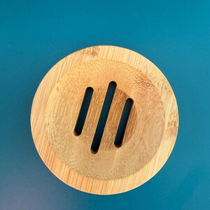 May include: A round, wooden soap dish with three drainage slots. The dish is made of light brown bamboo.