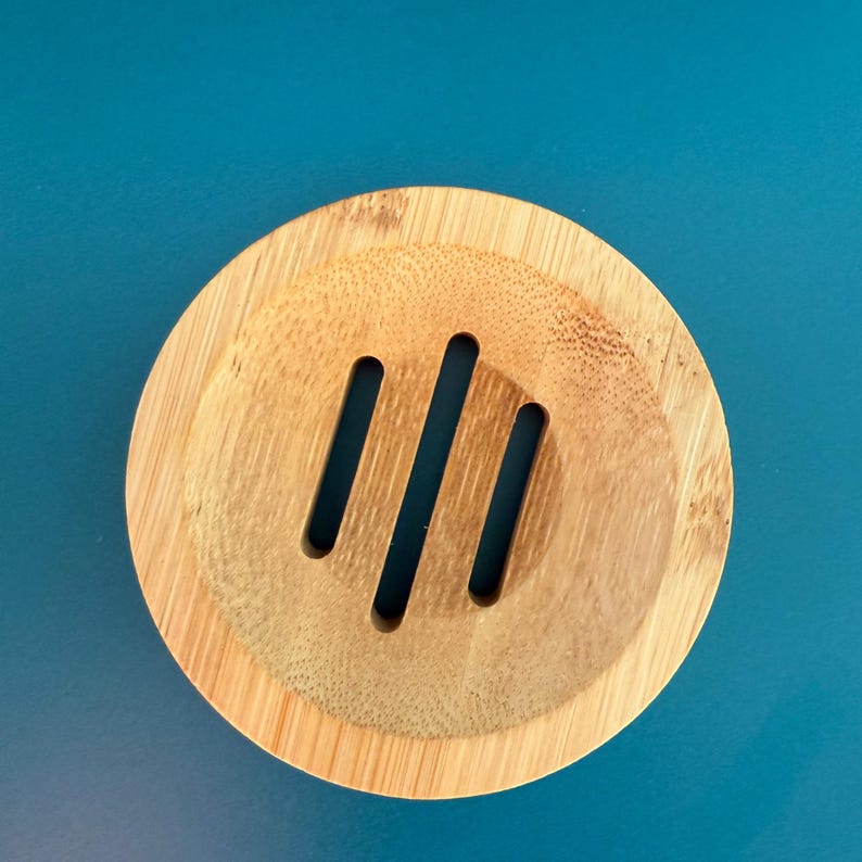 May include: A round, wooden soap dish with three drainage slots. The dish is made of light brown bamboo.