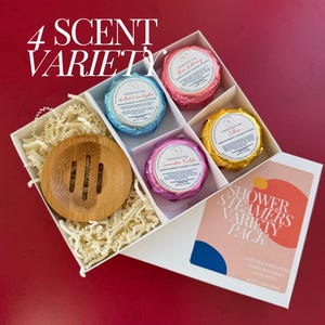 May include: A white box filled with four round shower steamers in a variety of colors and scents. The box also includes a small wooden soap dish. The box has a label that reads "Shower Steamers Variety Pack" and lists the scents: Lavender Fields, Coconut Milk, Mint & Eucalyptus, and Rose & Cherry.