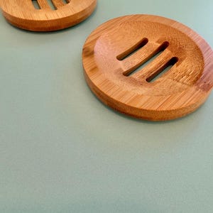 May include: Two wooden soap dishes with three drainage slots. The dishes are round and made of light brown wood. The bottom dish has three black rubber feet.