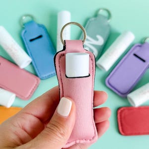 May include: A pink lip balm holder with a silver keyring, holding a white lip balm tube. Other colorful holders in blue, pink, purple, and red are visible. The holders are made of a soft material and have a slot for the lip balm.