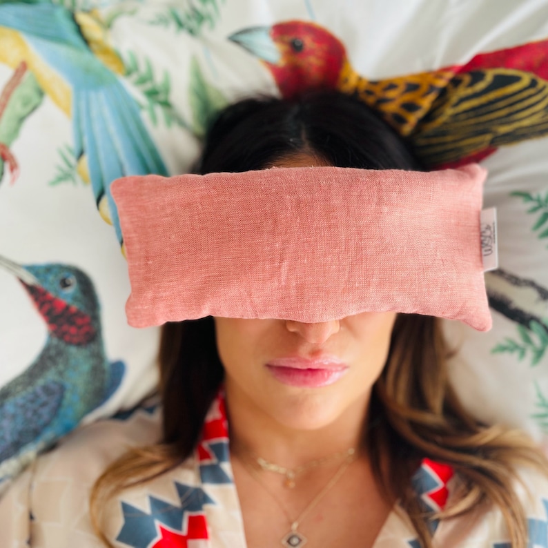 May include: A pink linen eye pillow with the word "Cozy" printed on it. The pillow is resting on a person's eyes, who is lying down on a bed with a floral patterned bedding.