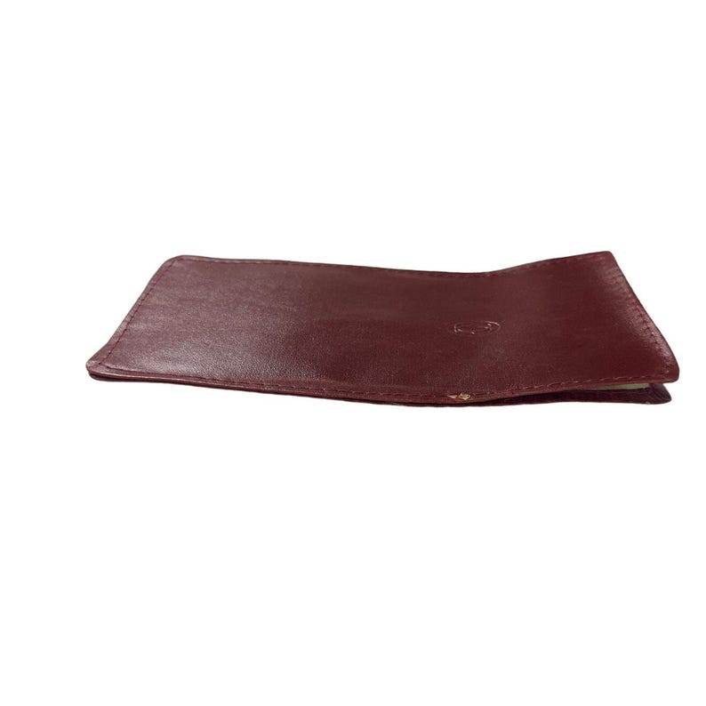 May include: A burgundy leather wallet lies on a white surface. The wallet is rectangular with slightly rounded corners.  It appears to be made of a smooth, supple leather. The color is a deep, rich burgundy. The wallet is closed, showing only the exterior.  It is a simple, elegant design suitable for everyday use. The leather shows subtle variations in tone and texture, suggesting a natural, high-quality material.