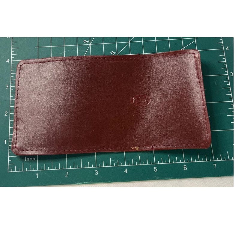 May include: A burgundy leather card holder is displayed on a green cutting mat.  The holder is rectangular with visible stitching along the edges.  A subtle logo is embossed on the front. The card holder measures approximately 4 inches by 6 inches.  It appears to be made of high-quality leather. The cutting mat has inch markings for measuring.