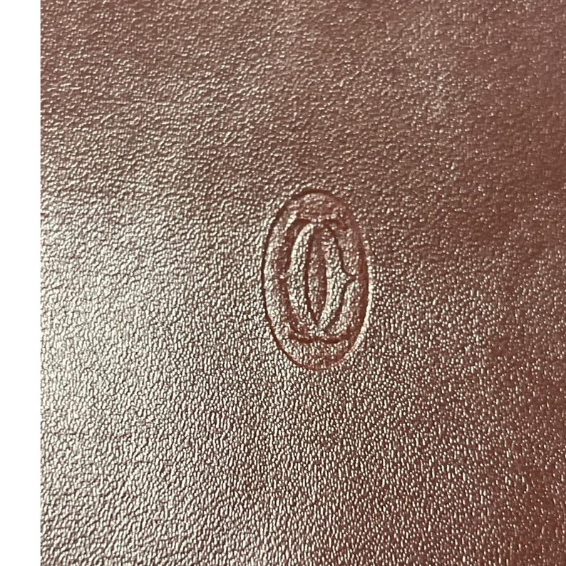 May include: Close-up view of a rose gold-colored textured surface with a subtle embossed Cartier logo. The logo is a circular emblem featuring the iconic Cartier lettering. The texture of the surface is fine and even, giving a luxurious feel. The color is a muted rose gold, with a slight sheen. The image highlights the brand's attention to detail and the high-quality materials used in their products.  The overall impression is one of elegance and sophistication, typical of the Cartier brand.
