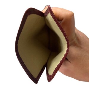 May include: Burgundy leather card holder case open to reveal a beige interior. The case is made of a soft, supple leather with a subtle texture.  The interior is a smooth, light beige fabric. The card holder is compact and designed to hold cards securely. The burgundy color is rich and deep, and the overall design is simple and elegant.