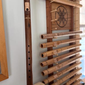 May include: A dark brown wooden flute with decorative accents and finger holes is held in front of a wall display of bamboo flutes. The display features multiple bamboo flutes mounted on a wooden frame, showcasing a variety of sizes and designs.