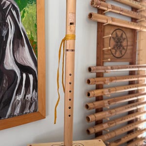 May include: A light-colored wooden flute with a yellow strap, featuring a series of finger holes and decorative bands. The flute is displayed on a wooden stand with an etched design. Other flutes are visible in the background.