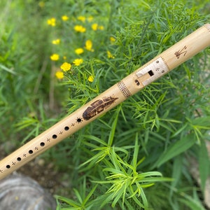 May include: A light-colored wooden flute with a feather design and decorative etchings. The flute has several finger holes and a mouthpiece. The background is blurred green foliage with yellow flowers.