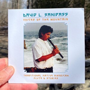 May include: A CD cover featuring the text "DAVID L. SANIPASS" and "VOICES OF THE MOUNTAIN." The cover art depicts a man playing a flute near the ocean. The text "TRADITIONAL NATIVE AMERICAN FLUTE & STORIES" is also present.