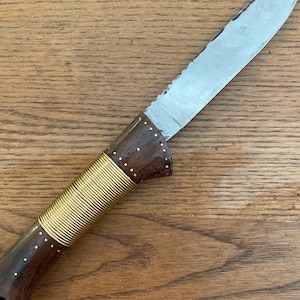 May include: A handmade knife with a wooden handle and a stainless steel blade. The handle is dark brown with brass accents and wrapped with gold-colored wire. The blade has a serrated edge.
