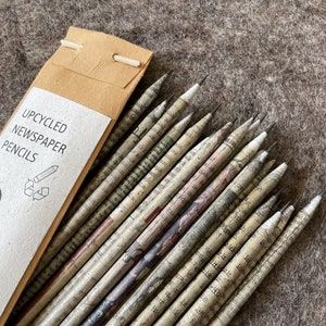 May include: A pack of five upcycled newspaper pencils. The pencils are made from recycled newspaper and have a natural, textured appearance. The pencils are packaged in a brown paper sleeve with the text "UPCYCLED NEWSPAPER PENCILS".