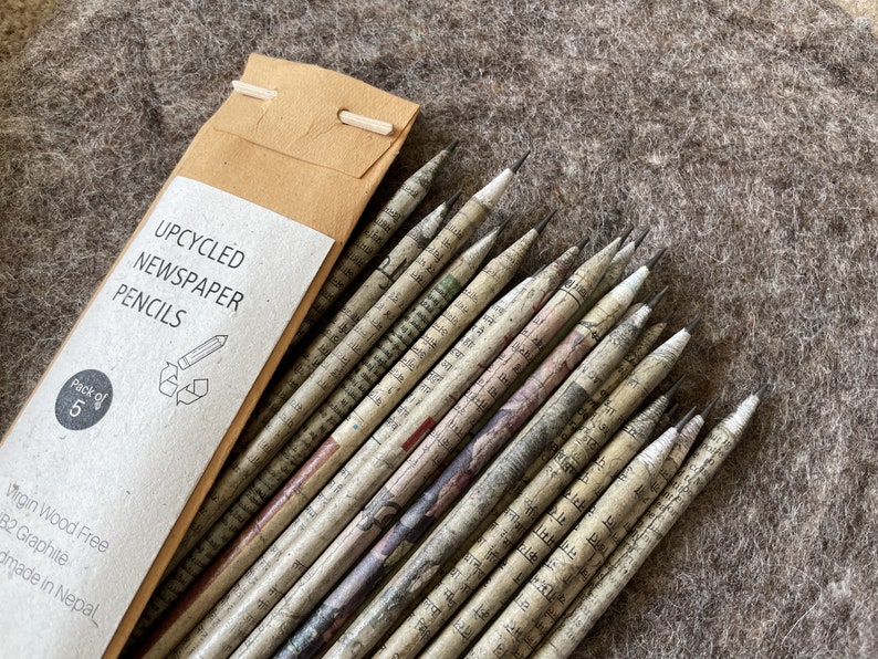 May include: A pack of five upcycled newspaper pencils. The pencils are made from recycled newspaper and have a natural, textured appearance. The pencils are packaged in a brown paper sleeve with the text "UPCYCLED NEWSPAPER PENCILS".