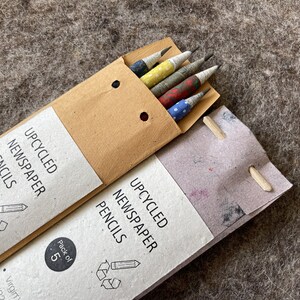 May include: Two packs of upcycled newspaper pencils. One pack is open, revealing five pencils with colorful patterned paper. The packaging reads "UPCYCLED NEWSPAPER PENCILS" and "Pack of 5". The pencils have a variety of patterns.