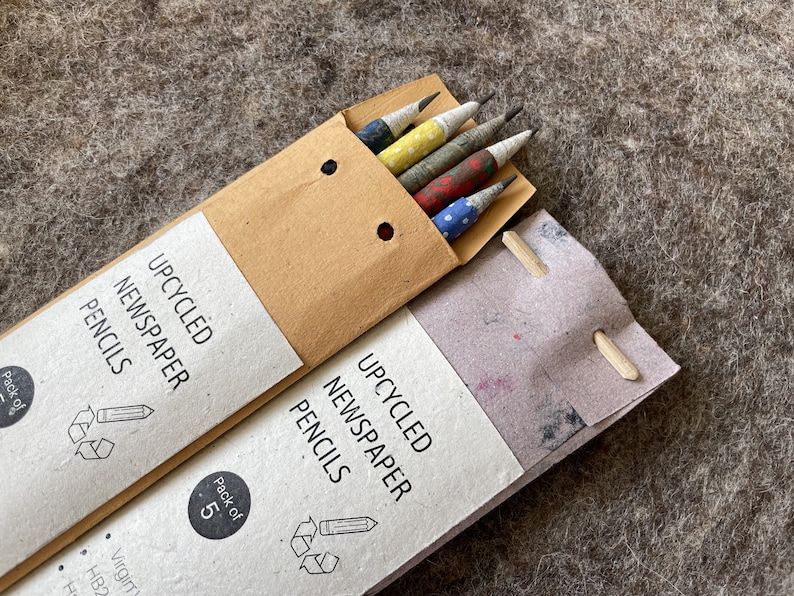 May include: Two packs of upcycled newspaper pencils. One pack is open, revealing five pencils with colorful patterned paper. The packaging reads "UPCYCLED NEWSPAPER PENCILS" and "Pack of 5". The pencils have a variety of patterns.