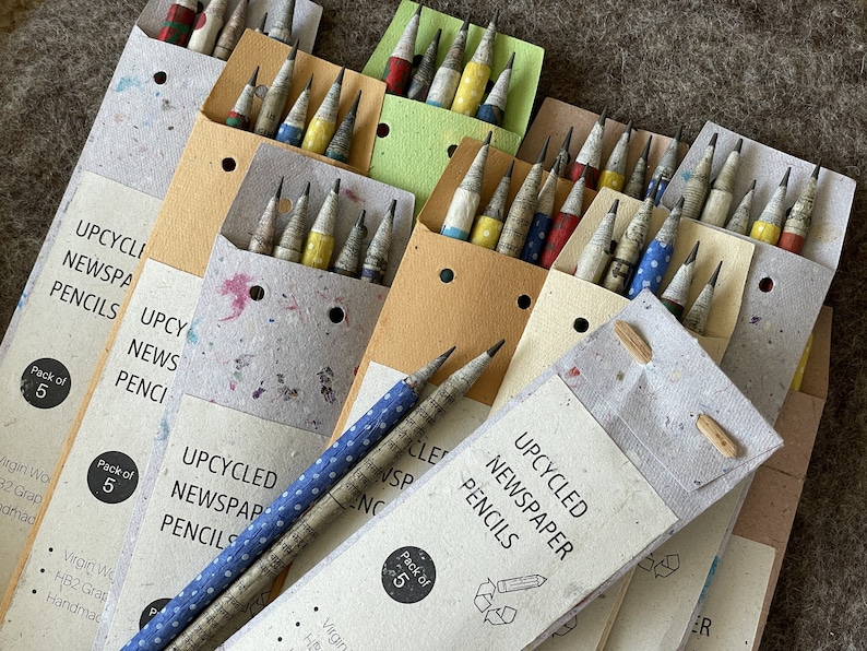 May include: A collection of upcycled newspaper pencils in packs of five. The pencils are made from recycled newspaper and have a unique, textured appearance. The packaging is made from recycled paper and features the text "UPCYCLED NEWSPAPER PENCILS".