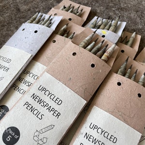 May include: A collection of upcycled newspaper pencils in packs of five. The pencils are made from recycled newspaper and are packaged in a light brown paper sleeve. The text "UPCYCLED NEWSPAPER PENCILS" is printed on the front of the sleeve.