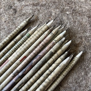 May include: A collection of pencils with a unique design. Each pencil is wrapped in paper printed with text and patterns. The pencils are arranged on a textured, neutral-colored surface, showcasing their distinct appearance.