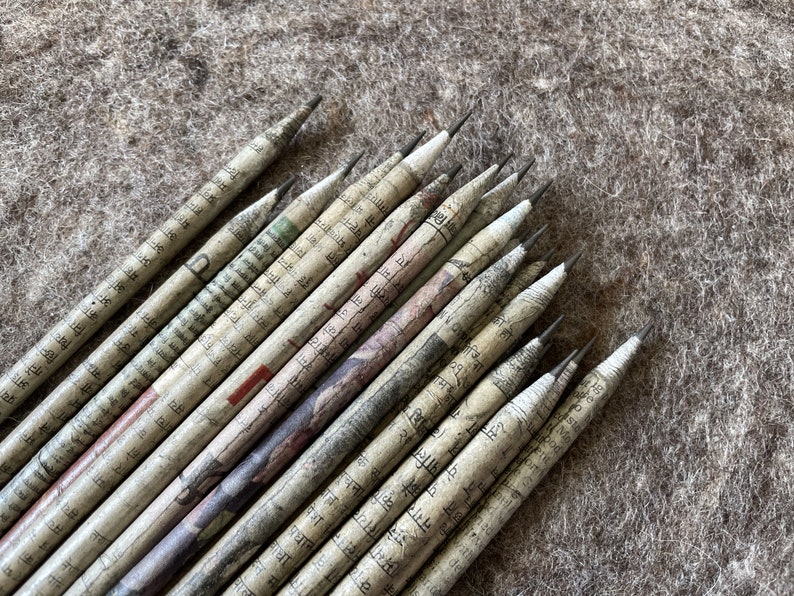 May include: A collection of pencils with a unique design. Each pencil is wrapped in paper printed with text and patterns. The pencils are arranged on a textured, neutral-colored surface, showcasing their distinct appearance.