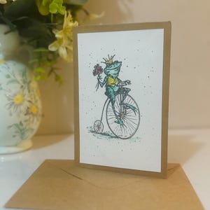 Carte de voeux aquarelle scintillante peinte à la main. Prince grenouille fantaisiste sur un monocycle avec des fleurs.