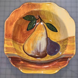May include: A ceramic serving bowl with a yellow, orange, and brown glaze. The bowl features a hand-painted design of a pear and a fig.