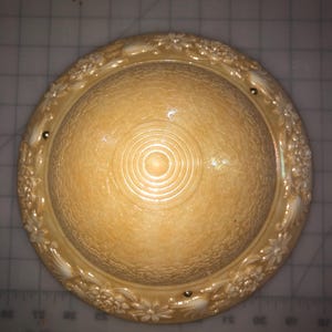 May include: A cream-colored, round decorative bowl with a textured surface and a floral border. The center of the bowl has concentric circles. Small gold-colored accents are placed around the rim.