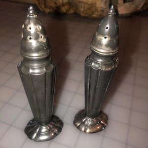 May include: Two antique silver salt and pepper shakers. Each shaker has a conical shape with vertical fluting and a decorative base. The tops have small holes for dispensing and a pointed finial. The silver has a tarnished appearance.