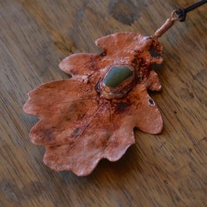 May include: A copper oak leaf pendant with a green stone set in the center. The pendant is hanging from a brown leather cord.