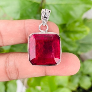 May include: A square-cut, deep red gemstone pendant set in a silver frame. The pendant features a faceted surface and a decorative silver bail at the top, designed for attachment to a necklace chain, showcasing a vibrant and translucent red hue.