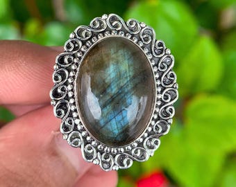 May include: Oval-shaped silver ring featuring a large, iridescent labradorite stone. The stone displays flashes of blue and green. The ring has an ornate, filigree-style setting with a detailed, swirling design around the stone.