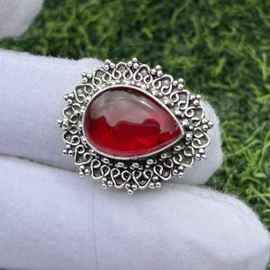 Peut inclure: Une bague argentée ornée d'une grosse pierre rouge foncé en forme de goutte d'eau. La pierre est sertie dans un entourage argenté ajouré et décoratif avec un motif floral. La bague est présentée sur une surface blanche sur fond vert.